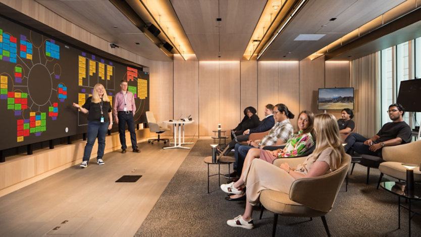 Two presenters stand at a large LED wall showing a design thinking note board as audience members sit engaged