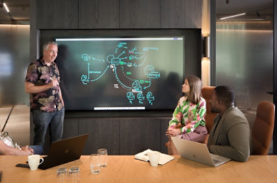 A person stands by a Surface Hub showing a whiteboard as a three others are seating nearby engaging in conversation
