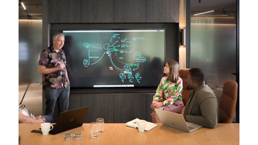 A person stands by a Surface Hub showing a whiteboard as a three others are seating nearby engaging in conversation