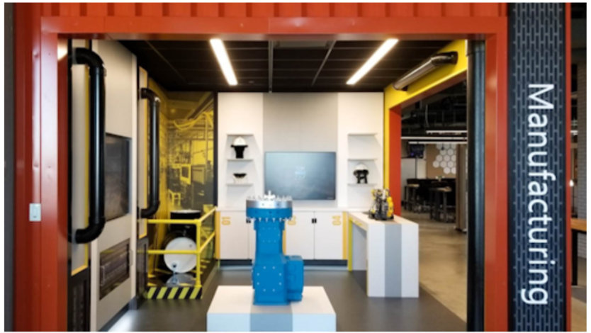 A red shipping container containing a themed experience area titled 'manufacturing'.  The floor holds a blue industrial pump and a shelf at the rear holds Hololens.