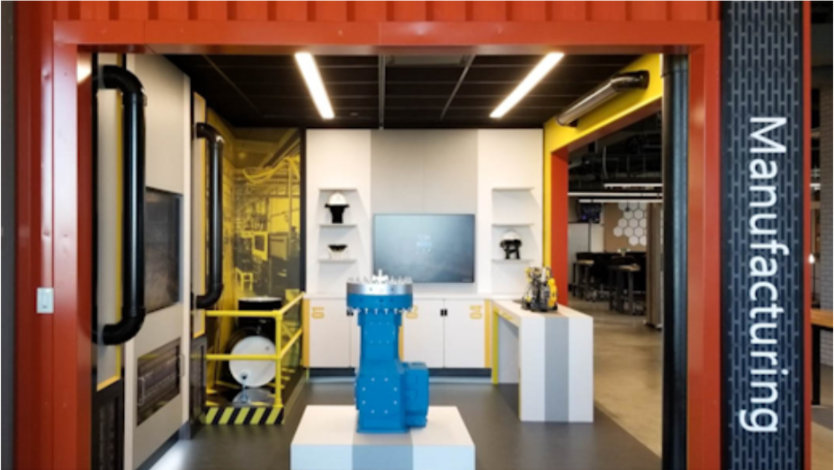 A red shipping container containing a themed experience area titled 'manufacturing'.  The floor holds a blue industrial pump and a shelf at the rear holds Hololens.