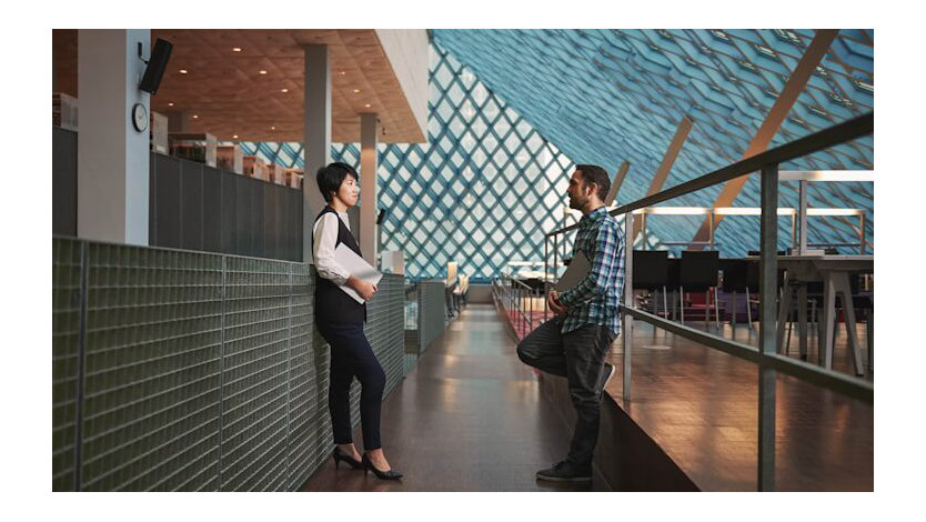 Two people talk together in a passageway in a large open area in a building.