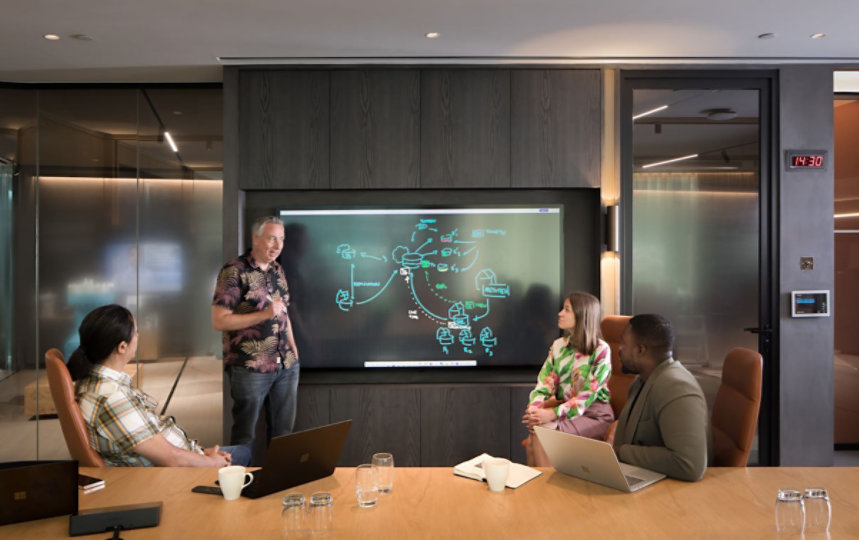 A person stands by a Surface Hub showing a whiteboard as a three others are seating nearby engaging in conversation