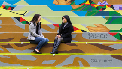 Two women sit outside on stairs while talking.