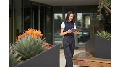 Woman in business attire looking at tablet, walking between planters outside of a building.