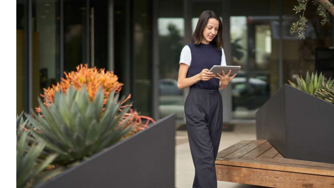 Woman in business attire looking at tablet, walking between planters outside of a building.