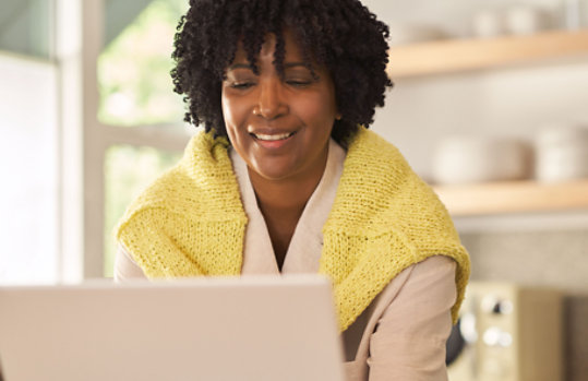 A smiling woman in a yellow sweater types on her laptop screen.