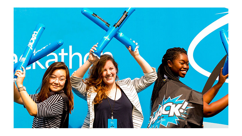 A group of three females celebrate Hackathon and are waving Hackathon swag in the air.