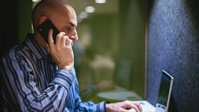 A man talks on a cell phone while working on a computer in an isolated booth.