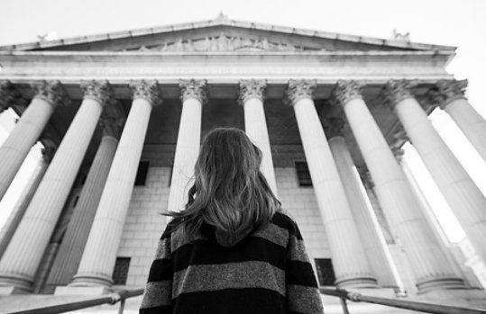 Black and white image of a woman looking up at a courthouse from the outside.