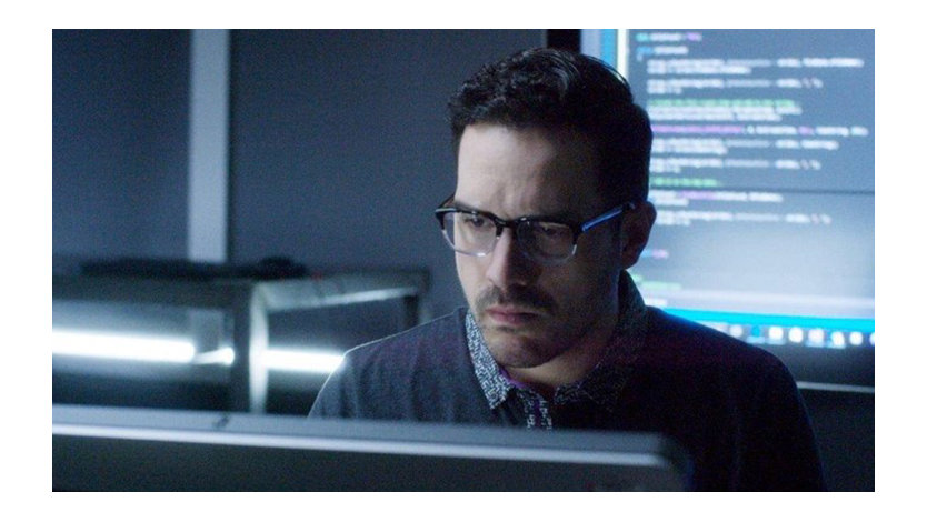 Nelson Palmer, do vídeo de treinamento da SBC, trabalhando em um computador em um escritório escuro, com uma tela atrás dele exibindo códigos de computador.