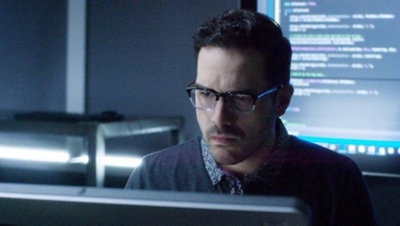 Nelson Palmer from the SBC training video working at a computer in a darkened office, with a screen behind him displaying computer code.