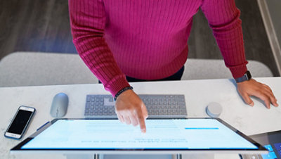 Top-down view of a man wearing a dark red shirt working on a Microsoft Surface Studio with a phone next to him on the desktop along with a mouse, keyboard, and Surface Dial.