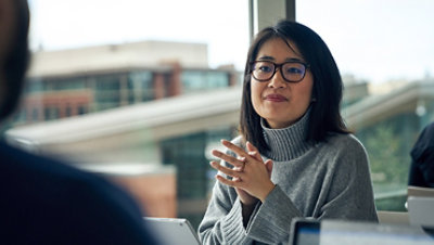 Woman with glasses sitting at a community table in a common area with her hands clasped together.