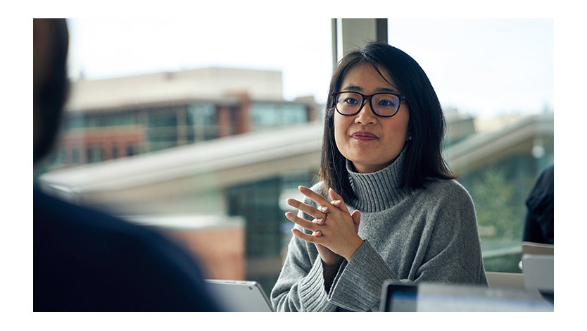 Woman with glasses sitting at a community table in a common area with her hands clasped together.