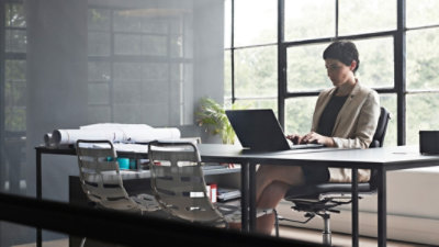 A woman is sitting at a long desk and typing on a laptop in a modern window office that has glass walls.