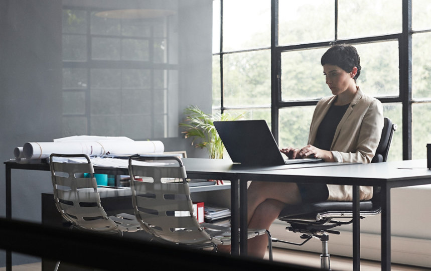 Uma mulher está sentada a uma mesa longa e digitando em um laptop em um escritório moderno com paredes de vidro. 