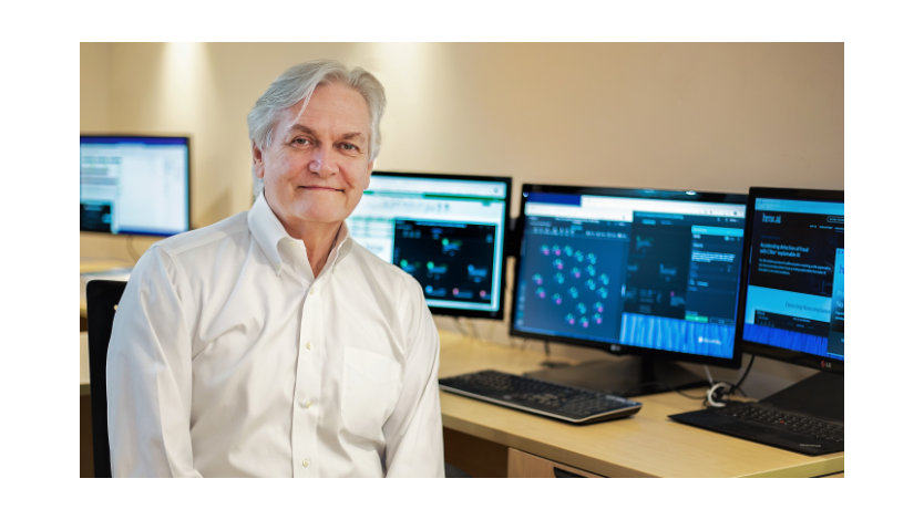 Michael Barnett smiles at the camera while sitting in front of his desk which shows multiple monitors that display an obscured background of data spreadsheets. 