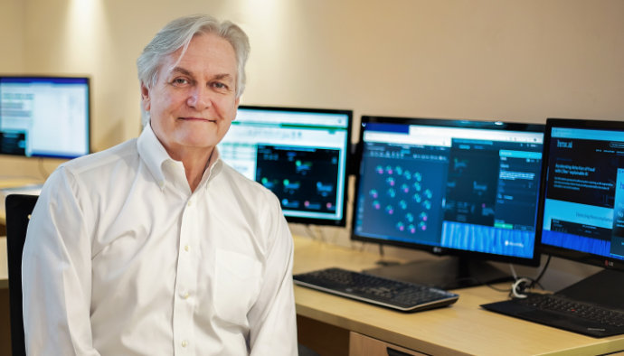 Michael Barnett smiles at the camera while sitting in front of his desk which shows multiple monitors that display an obscured background of data spreadsheets. 