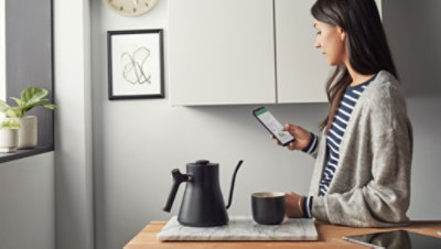 O persoană stând într-o bucătărie modernă, ținând o cană de cafea și uitându-se la un ecran de smartphone.