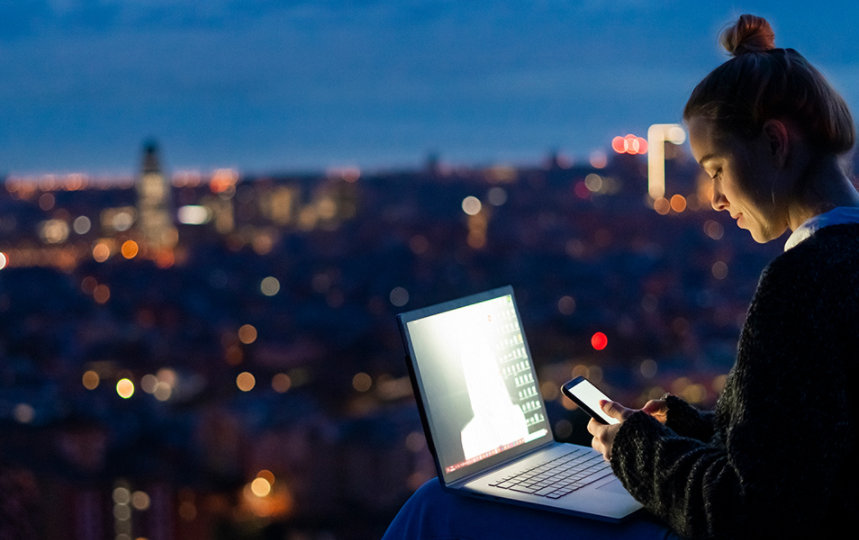 Woman using a cell phone and laptop at dawn overlooking a city.