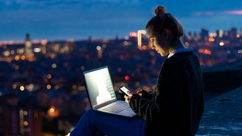도시가 내려다보이는 곳에서 새벽에 휴대폰과 노트북을 사용 중인 여성.