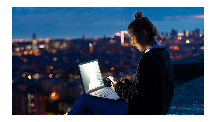 Woman using a cell phone and laptop at dawn overlooking a city.