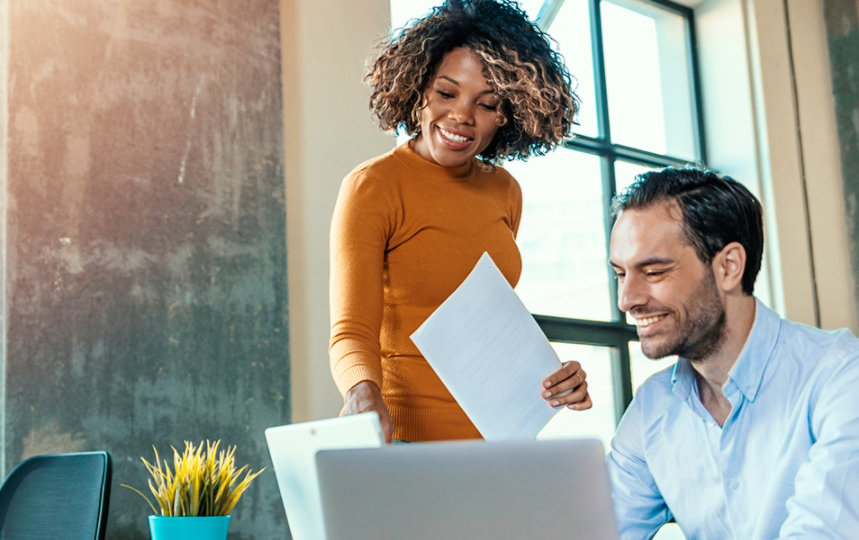 Woman standing in an office looks at a laptop screen with a seated male colleague.