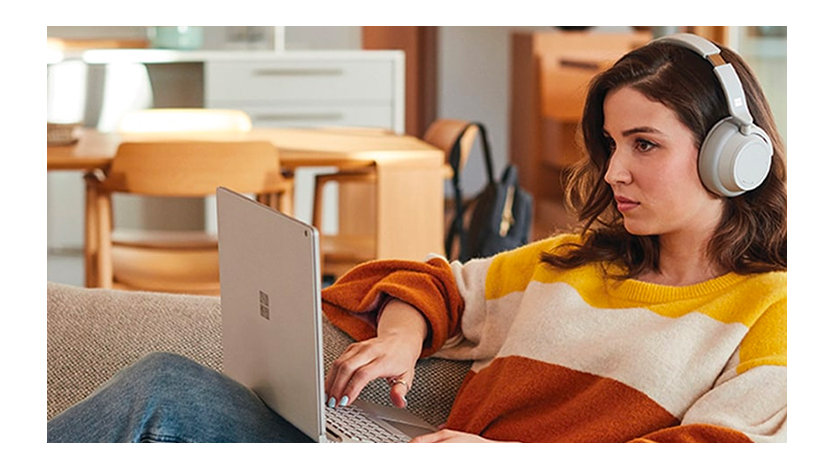 A woman uses a Surface laptop and Surface Headphones while lounging on a couch at home.