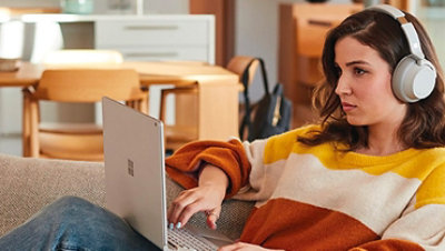 一名女子在家躺在沙發上使用 Surface Laptop 和 Surface Headphones。