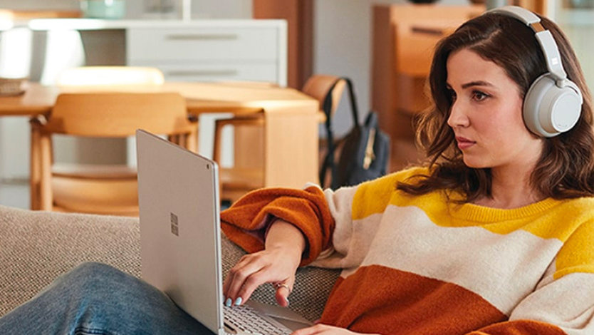 한 여성이 집안 소파에서 느긋한 자세로 Surface Laptop 및 Surface Headphones를 사용하는 모습.