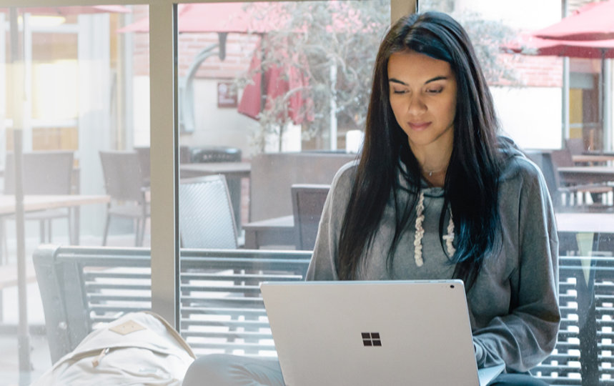Uma mulher jovem sentada com as pernas cruzadas em um parapeito usando um Surface Book no colo.