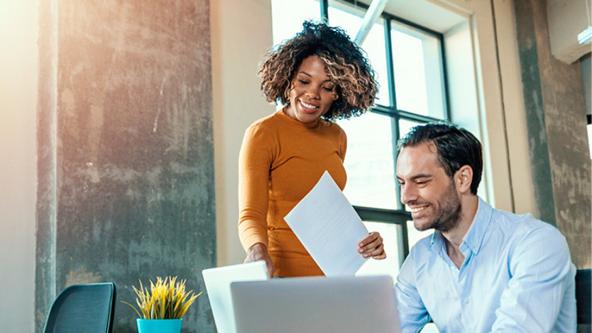 사무실에 서 있는 여성이 앉아 있는 남성 동료와 함께 노트북 화면을 바라보는 모습.