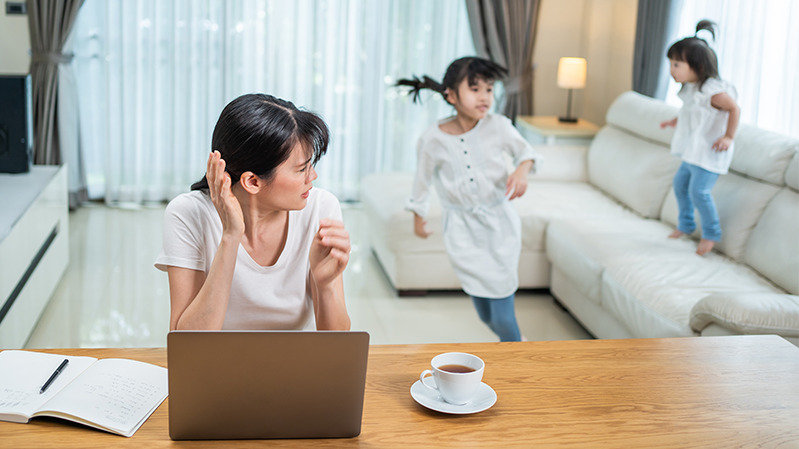 自宅のテーブルで仕事をしている女性と、後ろのソファで遊ぶ子供たち