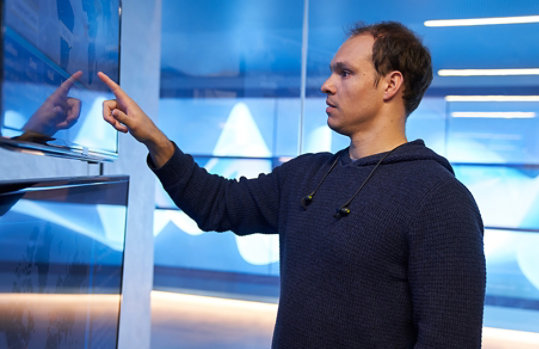 Man in a hooded sweater/sweatshirt inside a secure room, pointing at a geographic area displayed on a large monitor.