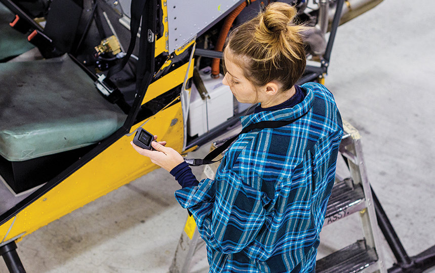 Overhead view of female worker standing in front of yellow helicopter at commercial manufacturing plant. She is holding an Askey IoT device; its screen displays rotor vibration levels. 