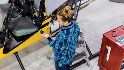 站在商业制造工厂黄色直升机前面的女性员工的俯视图。她拿着一台 Askey IoT 设备；屏幕上显示了旋翼的振动水平。