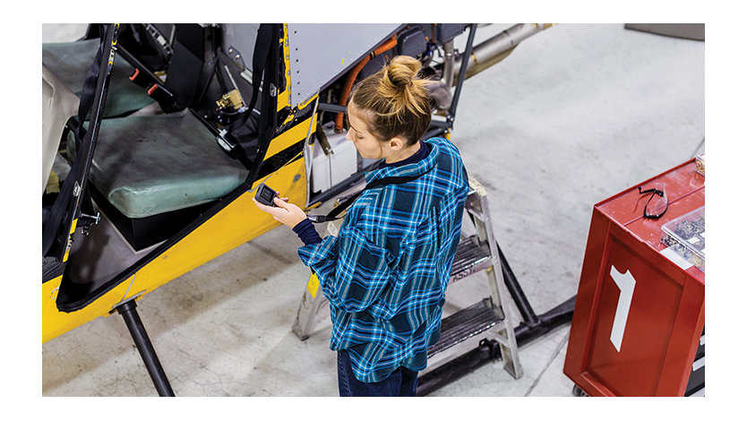 Overhead view of female worker standing in front of yellow helicopter at commercial manufacturing plant. She is holding an Askey IoT device; its screen displays rotor vibration levels.