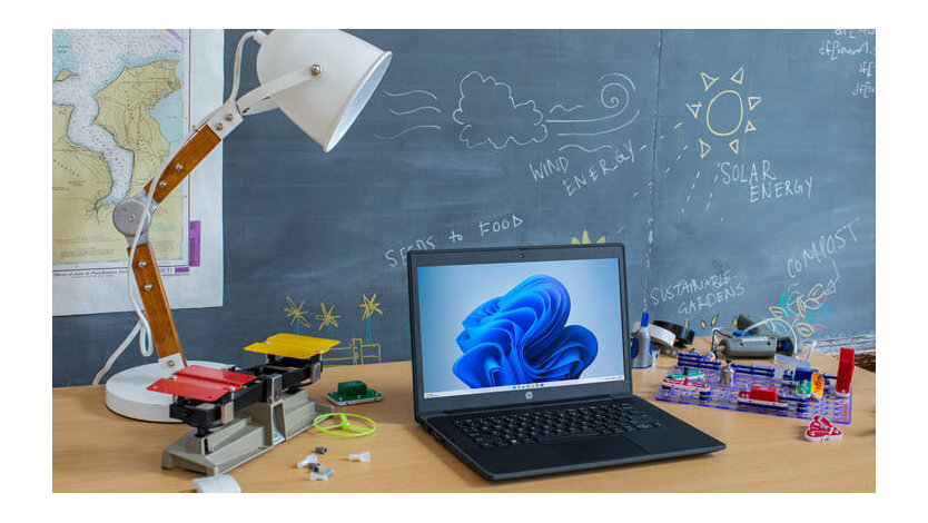 An open laptop sits on a table in a school classroom surrounded by science supplies with a blackboard in the background.