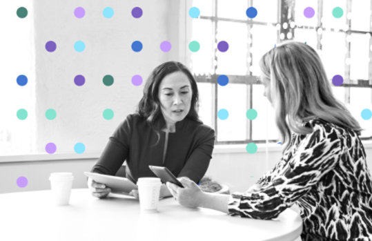 Two women sitting at a table in an office setting engaged in conversation. There is a layer of multi-colored dots on top of the image that are intended to represent data points. 