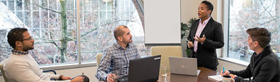 Four people in a meeting room, three are seated paying attention to the speaker, who is standing at the head of the table