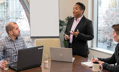 Osoba u odelu u sali za konferencije se predstavlja pred dvoje kolega koji sede za stolom na kojem se nalaze laptop računari, beležnice i šolja kafe.