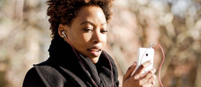 alt="Person outdoors, dressed warmly in a black scarf and coat, looking at a smartphone. Trees and a blurred background can be seen."