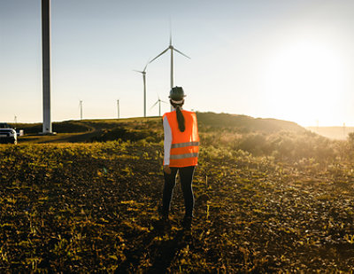 A woman in a reflective vest standing in a field with wind turbines.