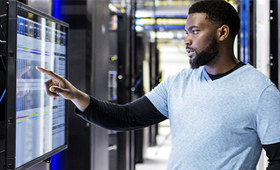 Un uomo in piedi davanti a un rack di server del data center, indica uno schermo su cui sono visualizzati dati.