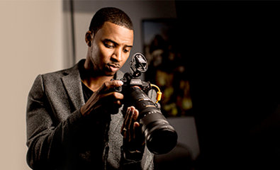 Muškarac u sivom sakou podešava kameru sa dugim objektivom u slabo osvetljenoj prostoriji.