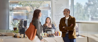 שלושה אנשים עומדים מול לוח מודעות מכוסה במגוון תמונות ודפים, מתעמקים בשיחה. 