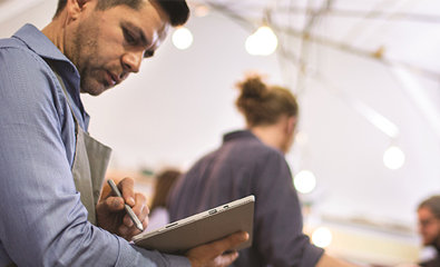 Un uomo con una maglia blu scrive su un quaderno mentre è in piedi. Altre persone sono presenti sullo sfondo.