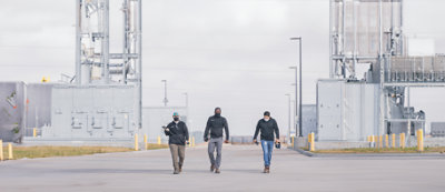 A man in a blue shirt writes in a notebook while standing. Other people are present in the background.