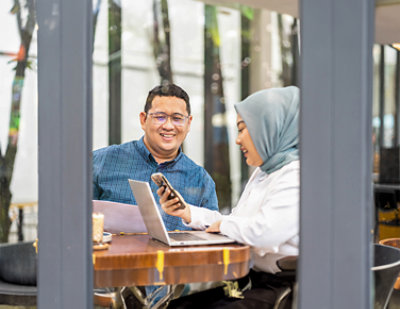 A woman wearing a head scarf holding a phone and looking at a laptop.
